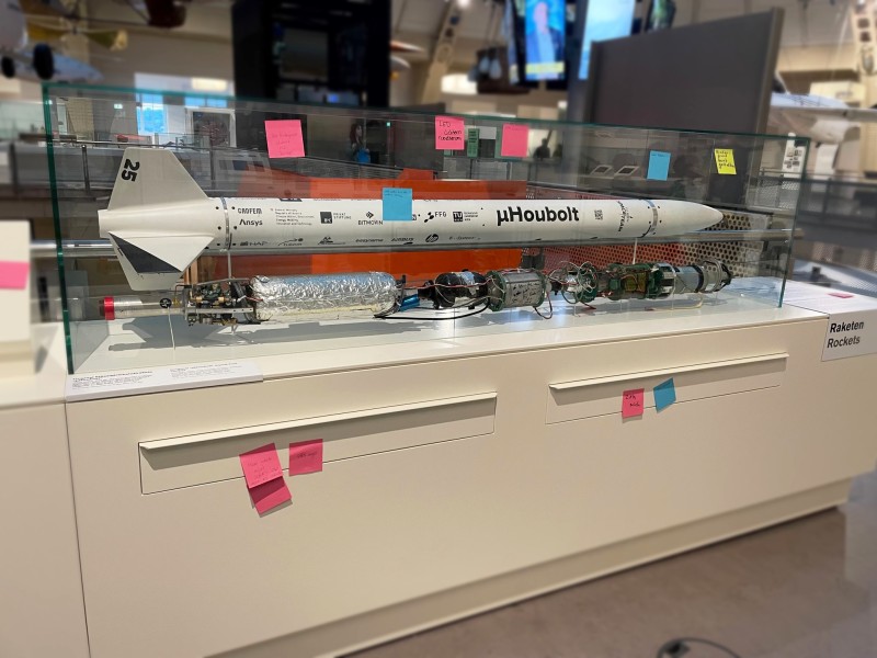 Ein Modell einer Rakete in einer Vitrine mit mehreren bunten Haftnotizen an der Vitrine und am Sockel, in einer Museumsausstellung mit der Aufschrift "Raketen.": Rückmeldungen zur und Vorschläge für die Raketenvitrine 