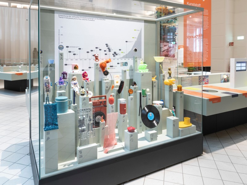 Vitrine aus der Ausstellung „Materialwelten“, die die Entwicklung von Kunststoffprodukten zeigt – mit bunten Alltagsgegenständen und einer Zeitleiste zur Produktionsmenge.: 