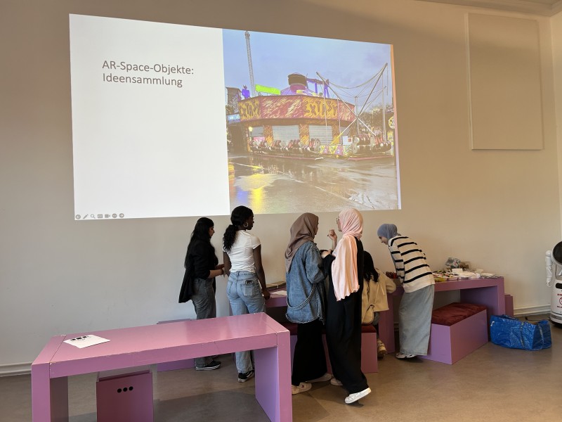 Gruppe Jugendlicher sammelt im Workshop Ideen zu AR-Objekten. Eine Präsentation mit Jahrmarkt-Foto und dem Titel „AR-Space-Objekte: Ideensammlung“ ist im Hintergrund projiziert.: Schwierige Entscheidungen stehen bevor: Welche Ideen will die Mission Control für das Design des AR-Objektes für die Mission Prater umsetzen?