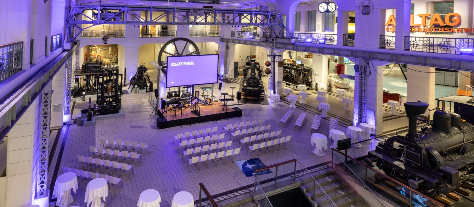 Festlich beleuchteter Mittelhalle des Technischen Museums Wien mit Bühne, Stuhlreihen und Stehtischen zwischen historischen Exponaten wie Dampfloks und Maschinen.: 