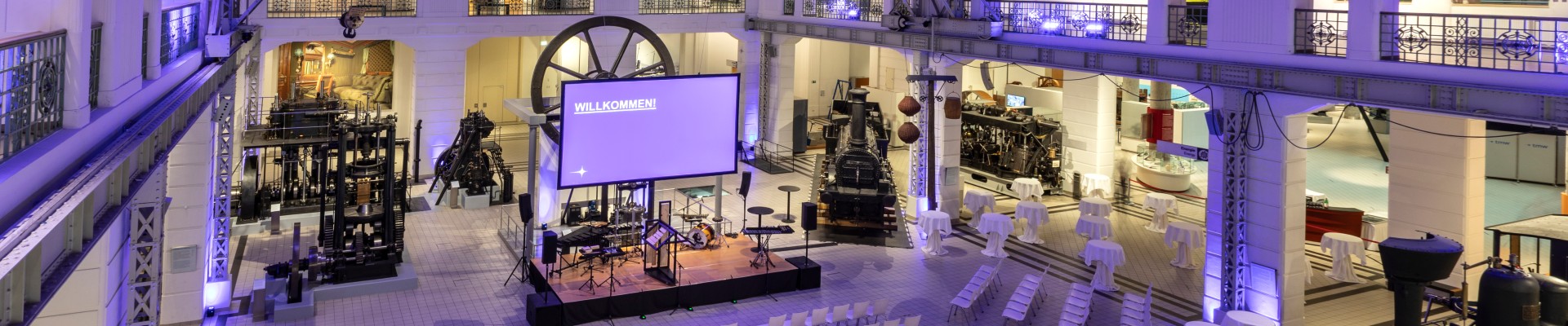 Festlich beleuchteter Mittelhalle des Technischen Museums Wien mit Bühne, Stuhlreihen und Stehtischen zwischen historischen Exponaten wie Dampfloks und Maschinen.: 