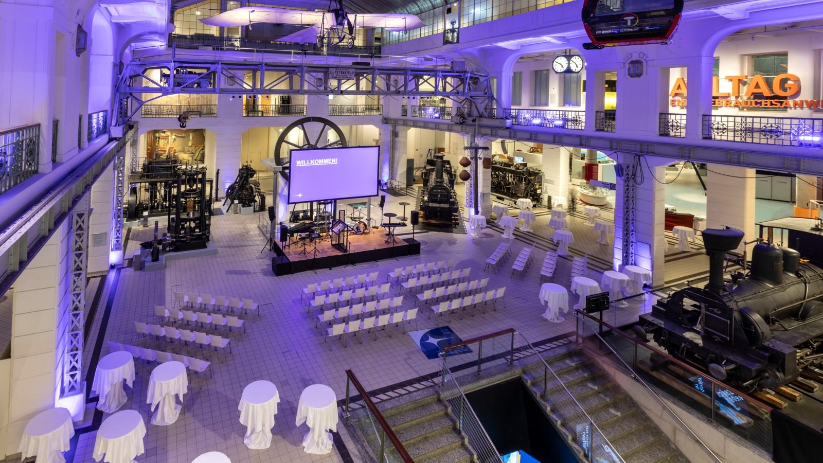 Festlich beleuchteter Mittelhalle des Technischen Museums Wien mit Bühne, Stuhlreihen und Stehtischen zwischen historischen Exponaten wie Dampfloks und Maschinen.: 