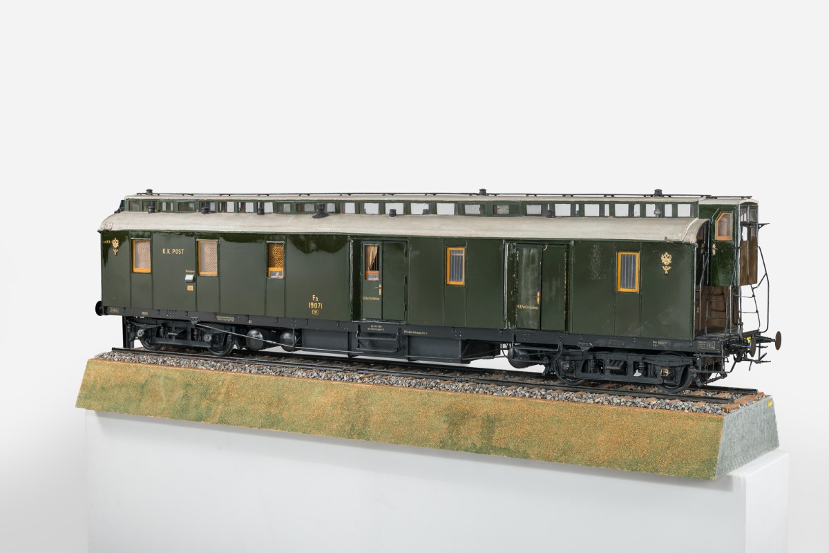 Vierachsiger Bahnpostwagen als Modell auf Gleisbett; langgestreckter, dunkelgrüner Wagen mit vielen Fenstern, Dachaufbauten und Türen, beschriftet „K.K. Post“, mit fein ausgearbeiteten Fahrwerksdetails.: Vierachsiger Bahnpostwagen (Modell, um 1914–1920; Maßstab: 1:10, Hersteller: Karl Löbl)