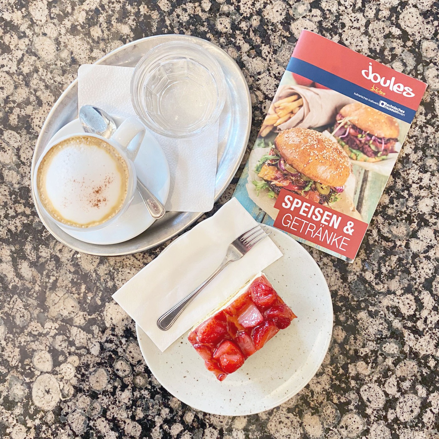 A cup of cappuccino, a glass of water, a strawberry cake, and a menu on a speckled granite table.: 