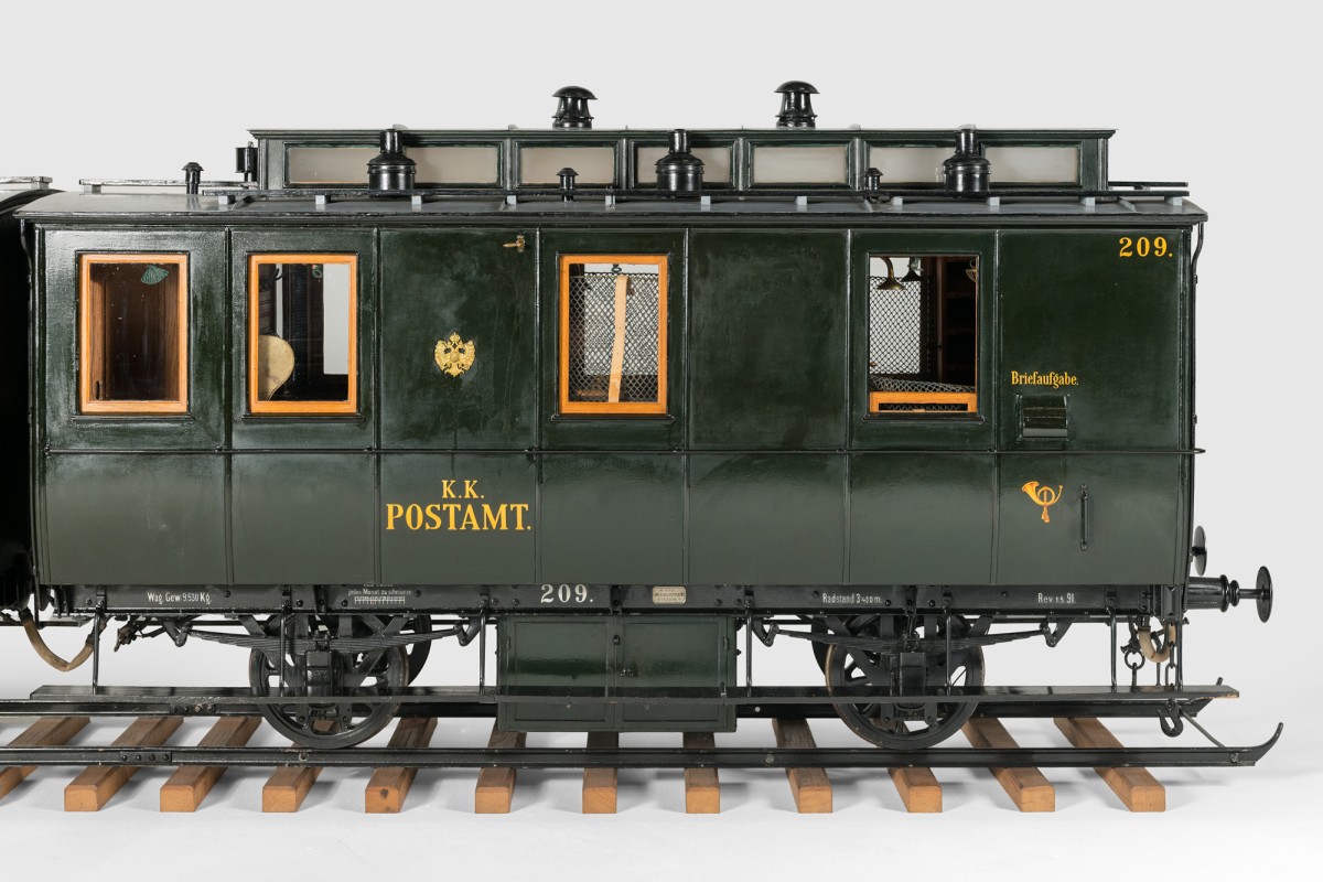 Seitenansicht eines zweiachsigen Bahnpostwagens als detailreiches Modell; dunkelgrüne Karosserie mit goldener Aufschrift „K.K. Postamt“, vergitterten Fenstern, Briefschlitz („Briefaufgabe“) und sichtbarem Fahrgestell auf Gleisen.: Zweiteiliger Bahnpostwagen (Modell, um 1900; Maßstab: 1:5), der Bildausschnitt zeigt den Kartierraum von außen mit vergitterten Fenstern
