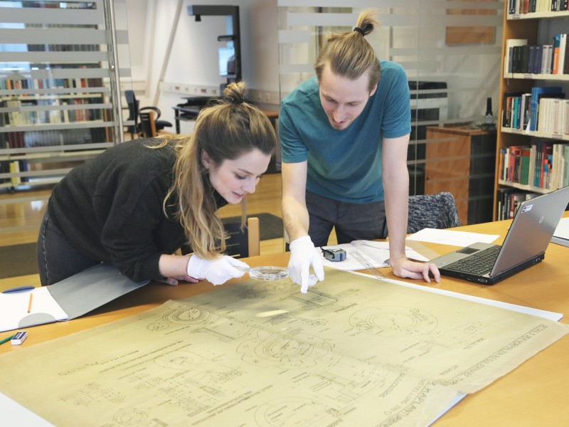 : A woman and a man study an old architectural plan of a building with a magnifying glass in the library