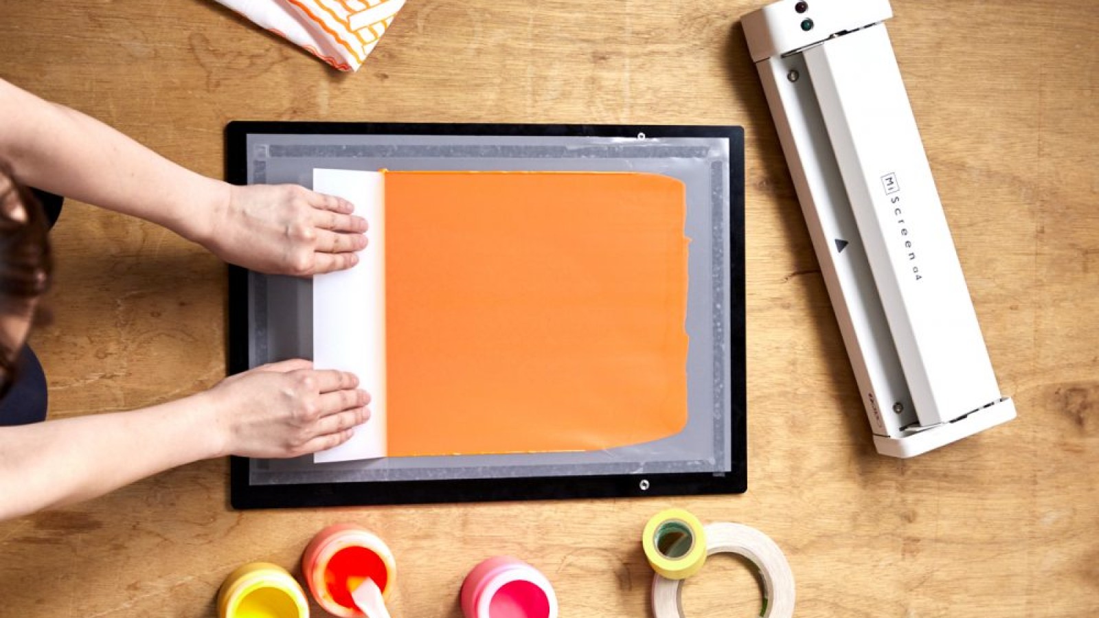 Hände drücken ein weißes Blatt auf einen orangenen Farbauftrag in einem Siebdruckrahmen; daneben liegen eine weiße Siebdruckmaschine, Farbbehälter, Klebebänder und ein Tuch auf einer Holzoberfläche.: 