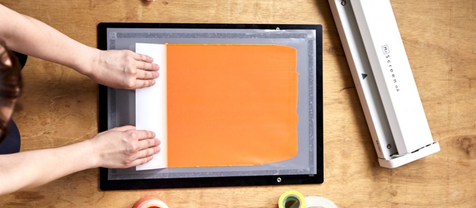 Hände drücken ein weißes Blatt auf einen orangenen Farbauftrag in einem Siebdruckrahmen; daneben liegen eine weiße Siebdruckmaschine, Farbbehälter, Klebebänder und ein Tuch auf einer Holzoberfläche.: 