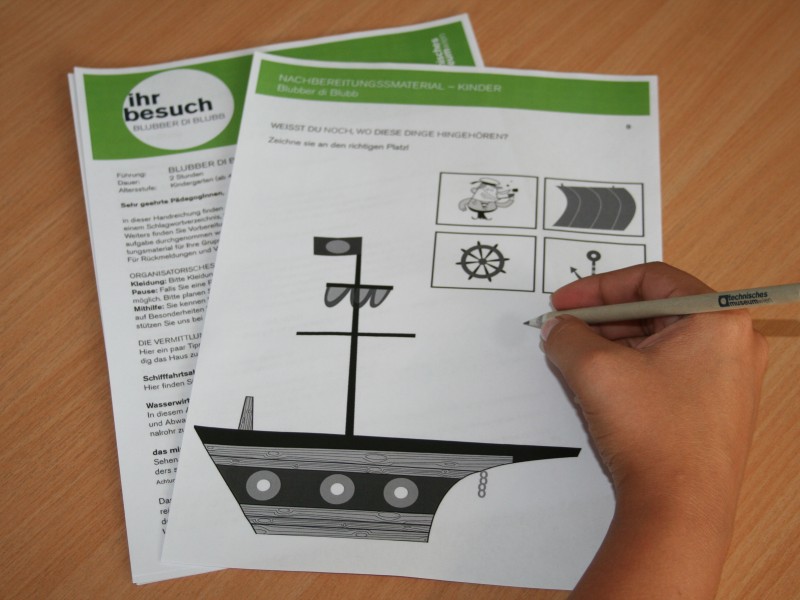 : Kinderhand beim Ausfüllen eines Papierbogens, auf dem die Illustration eines Schiffes abgebildet ist.