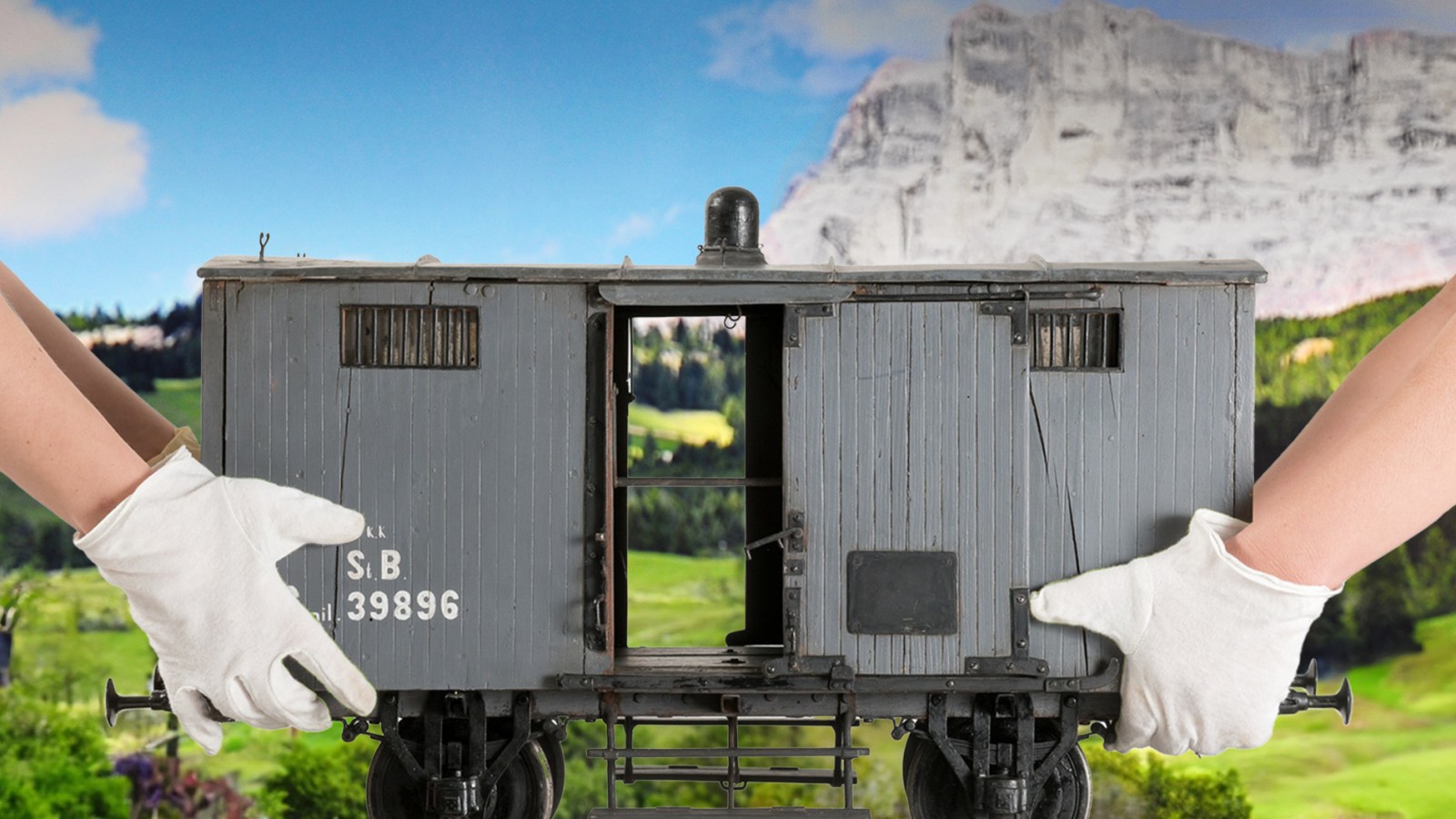 Zwei Hände mit weißen Handschuhen halten von links und rechts einen grauen Modell-Güterwagon mit der Aufschrift „S.B. 39896“ vor einer Landschaft mit grünen Hügeln und einem Felsenmassiv im Hintergrund.: