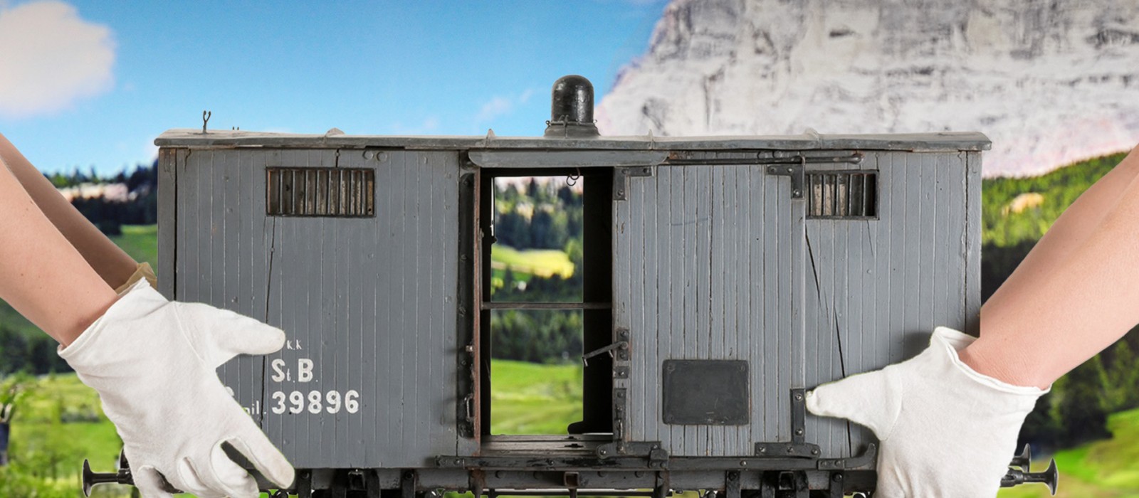 Zwei Hände mit weißen Handschuhen halten von links und rechts einen grauen Modell-Güterwagon mit der Aufschrift „S.B. 39896“ vor einer Landschaft mit grünen Hügeln und einem Felsenmassiv im Hintergrund.: