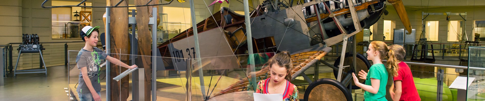 Vier Kinder sehen sich in der Ausstellung "Mobilität" das Flugzeug Aviatik (Berg) DI (101.37) an. Eines von ihnen füllt die Rätselrallye dazu aus. : 