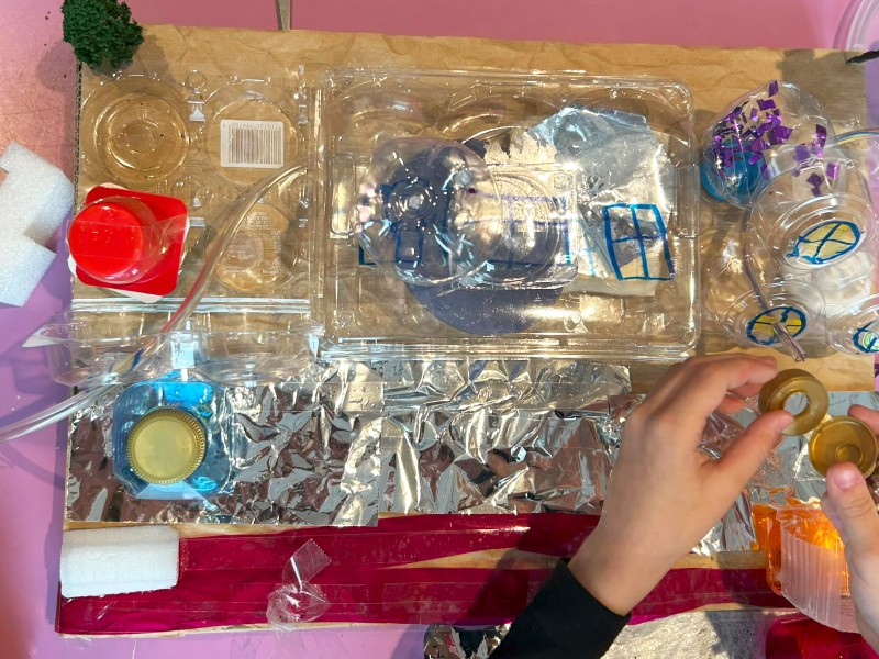 Children's hands working on a creative project using recycled materials, including plastic containers, bottle caps, foil, and colored tape.: Enthusiastic building and creating for the 'MISSION AT HOME'.