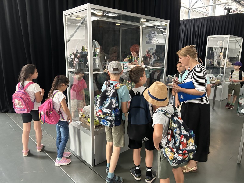 Kindergruppe mit Begleitperson betrachtet eine Vitrine mit Alltagsobjekten; Ausstellungssituation mit mehreren Vitrinen im Hintergrund.: Die jungen Architekt:innen bestaunen nochmals ihre Habitate und die ihrer Klassenkolleg:innen.