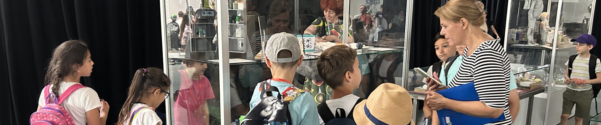 Kindergruppe mit Begleitperson betrachtet eine Vitrine mit Alltagsobjekten; Ausstellungssituation mit mehreren Vitrinen im Hintergrund.: Die jungen Architekt:innen bestaunen nochmals ihre Habitate und die ihrer Klassenkolleg:innen.