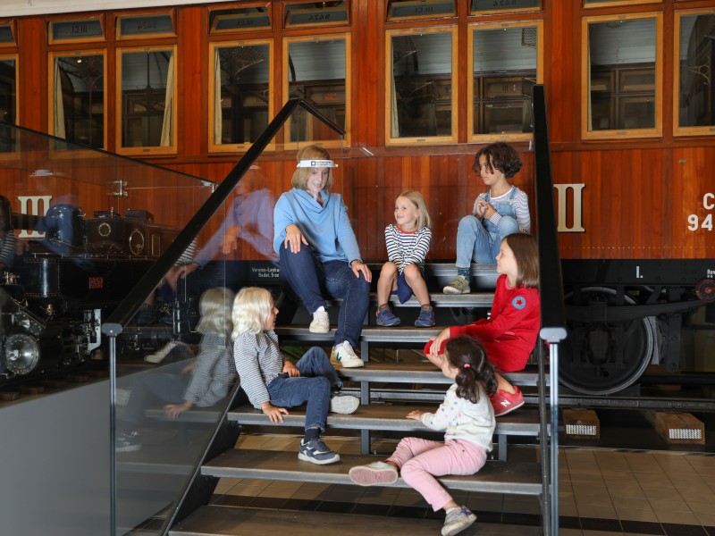 : Children sit together with a tour guide on the stairs from Sisi´s Hofsalonwagen. It is part of the exhibition "Lok.Erlebnis" 