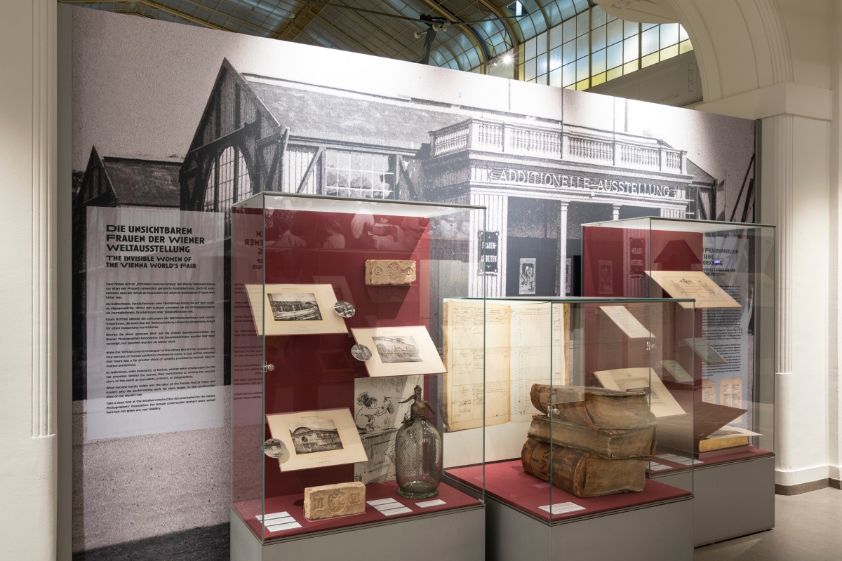 Vitrinen der Ausstellung „Women at Work“, im Hintergrund eine Wand mit dem Bild der „Additionellen Ausstellung“, in der der „Frauen-pavillon“ der Weltausstellung untergebracht war