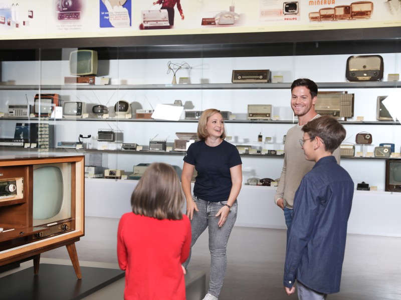 Eine Kulturvermittlerin mit einer Familie in der Ausstellung medien.welten, im Hintergrund historische Fernsehgeräte: 