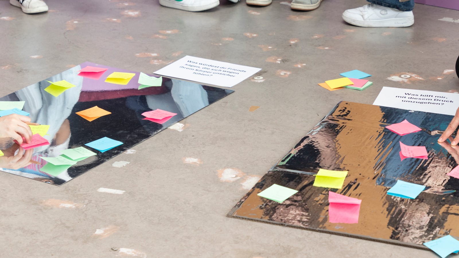 Teilnehmer setzen Post-It-Notizen auf Plakate mit Fragen zum Thema Körperbild und Stressbewältigung.: