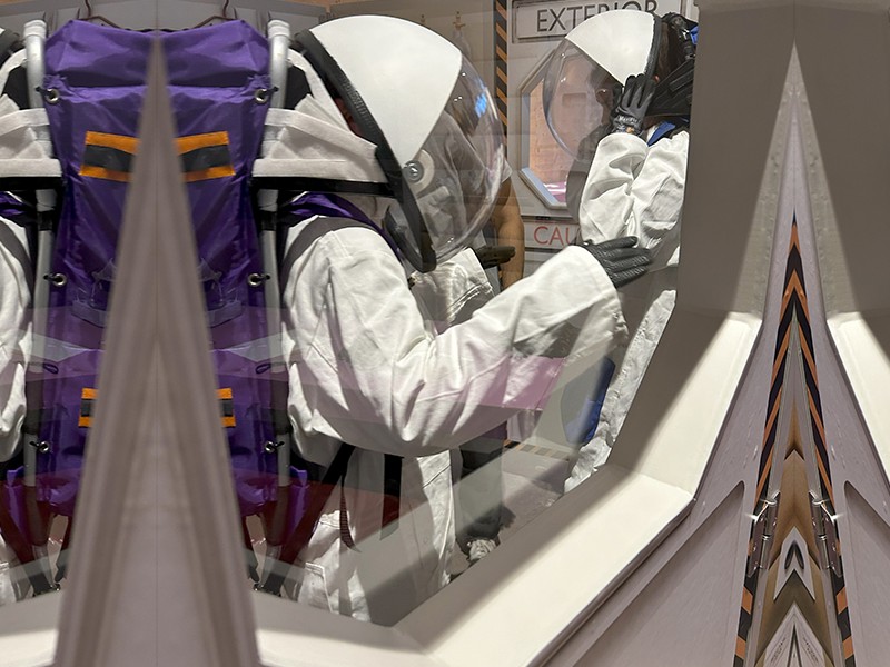 Zwei Personen in weißen Astronautenanzügen und Helmen sind durch ein Fenster zu sehen, wie sie in einer simulierten Schleuse mit den Aufschriften "EXTERIOR" und "CAUTION" stehen.: 