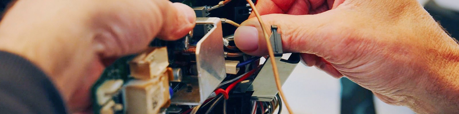 Nahaufnahme der Hände einer Person, die konzentriert an der offenliegenden Elektronik und Verkabelung eines Geräts arbeitet. Man sieht filigrane Drähte, Stecker und Bauteile, während die Finger präzise Handgriffe ausführen.