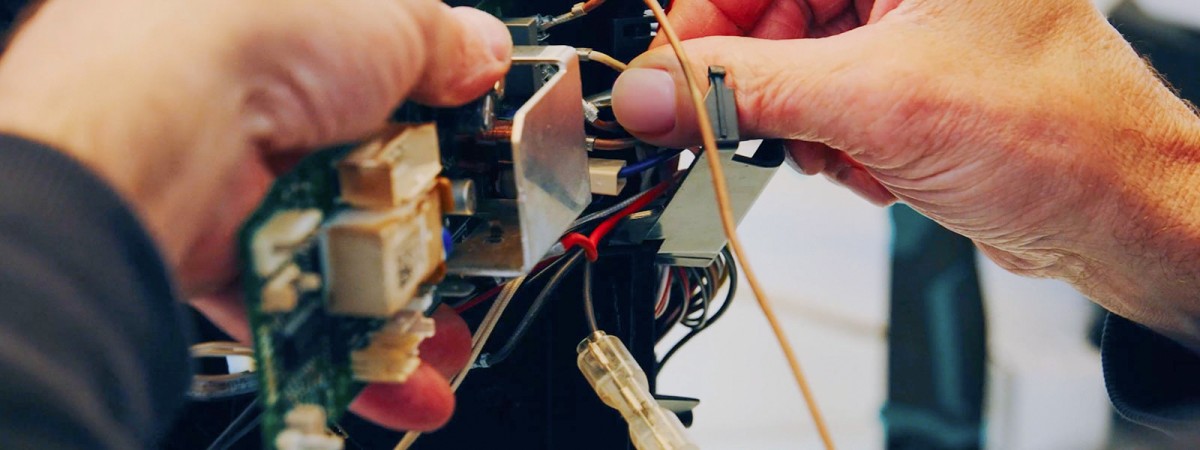 Nahaufnahme der Hände einer Person, die konzentriert an der offenliegenden Elektronik und Verkabelung eines Geräts arbeitet. Man sieht filigrane Drähte, Stecker und Bauteile, während die Finger präzise Handgriffe ausführen.
