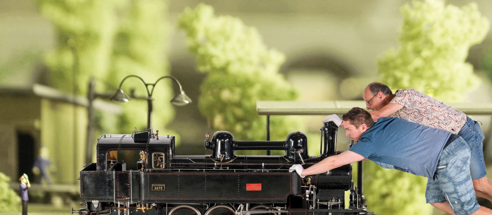 Zwei erwachsene Personen lehnen sich nach vorne und drücken eine große schwarze Modelllokomotive auf einem Gleis; im Hintergrund stehen modellhafte Bäume und Gebäude.: 