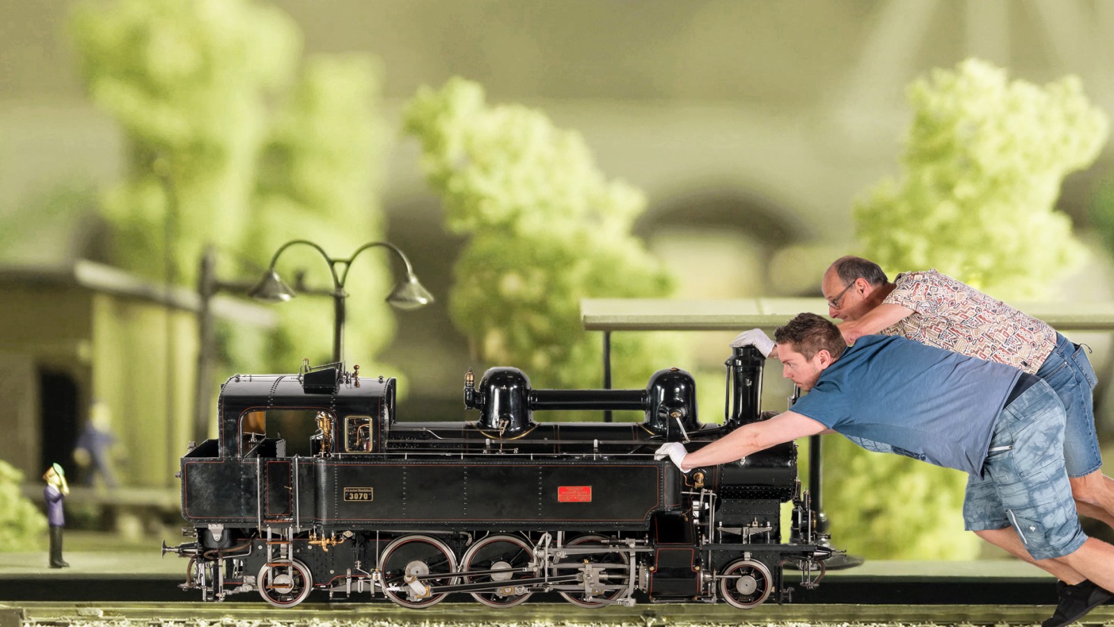Zwei erwachsene Personen lehnen sich nach vorne und drücken eine große schwarze Modelllokomotive auf einem Gleis; im Hintergrund stehen modellhafte Bäume und Gebäude.: 