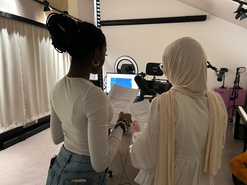 Zwei junge Frauen stehen in einem Tonstudio vor einem Mikrofon und lesen gemeinsam von einem Skript. Im Hintergrund sind technische Geräte und ein Bildschirm sichtbar.: Konzentration ist angesagt: Die Mission Control hat aber schon Erfahrung mit Tonaufnahmen und daher alles im Griff.