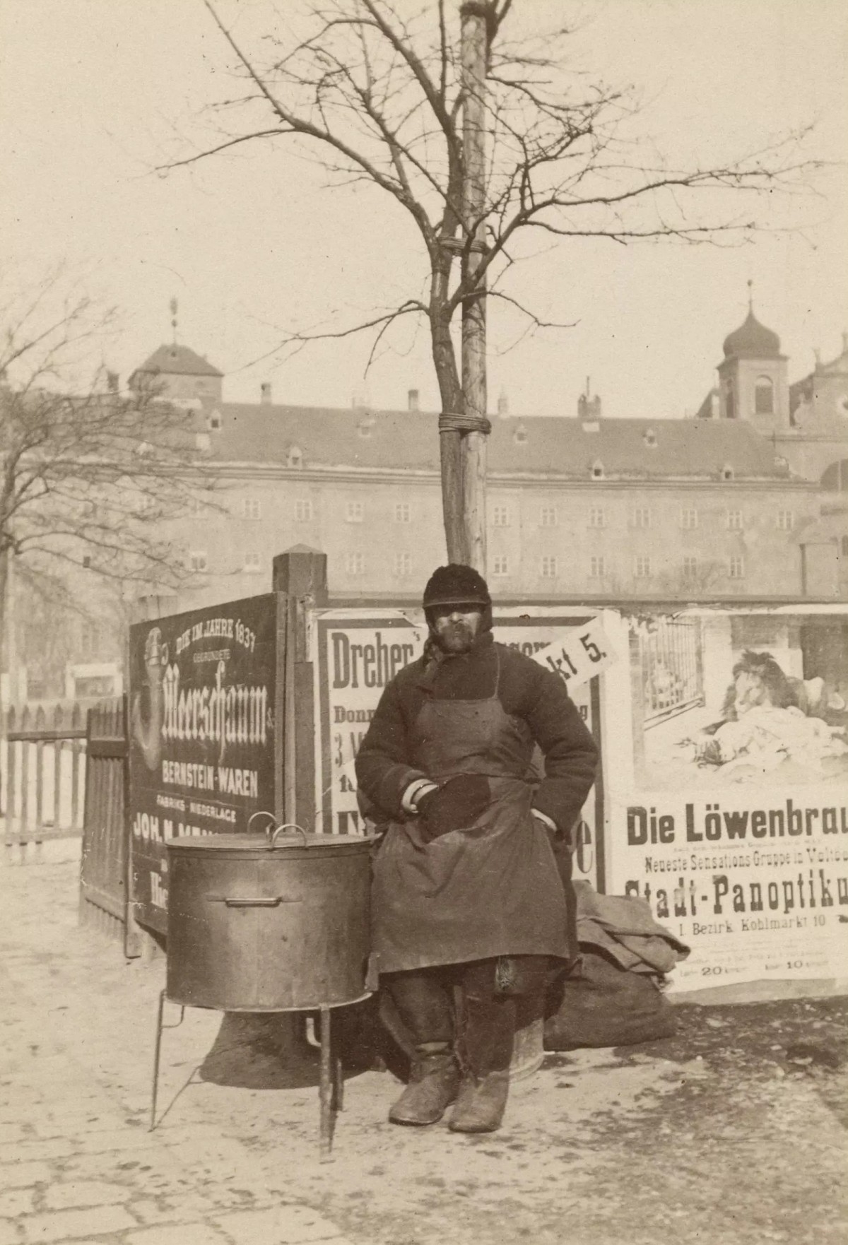 Historisches Foto eines Mannes in Winterkleidung und Schürze, der neben einem metallenen Maroniofen steht und seine Ware anbietet. Im Hintergrund sind Werbeflächen mit Texten wie „Meerschaum“ und „Die Löwenbrauerei“ sowie ein großes Gebäude mit Türmen zu sehen: ... das Straßenbild in Wien