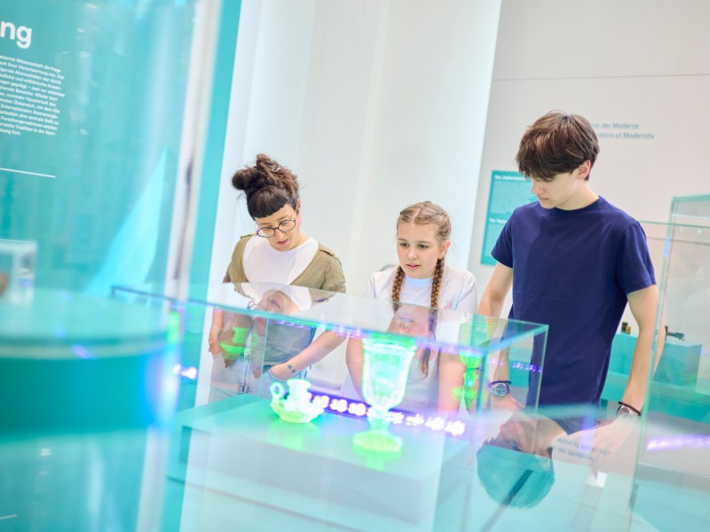 Drei Personen, eine erwachsene Frau und zwei Kinder, betrachten in einem Museum aufmerksam grün leuchtende Objekte in einer Glasvitrine: