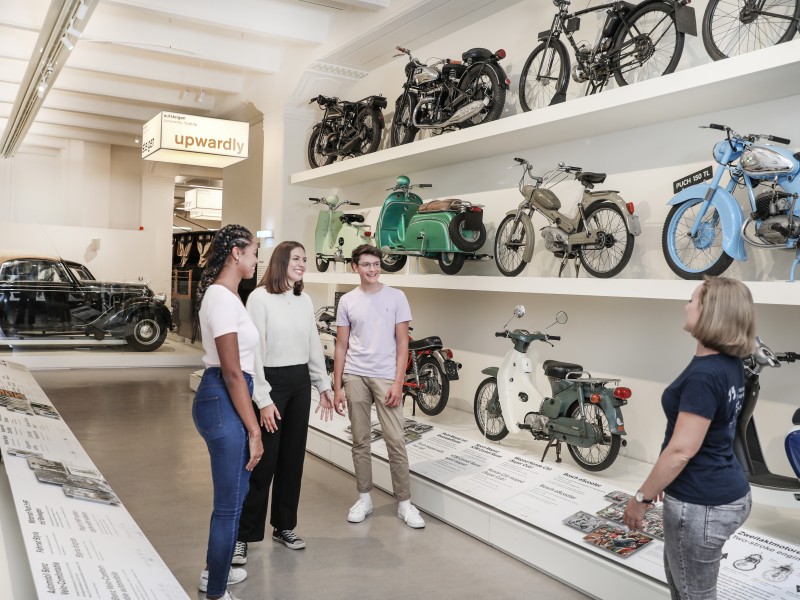 : A small group of teenagers look at various mopeds and motorcycles in the exhibition "Mobility".