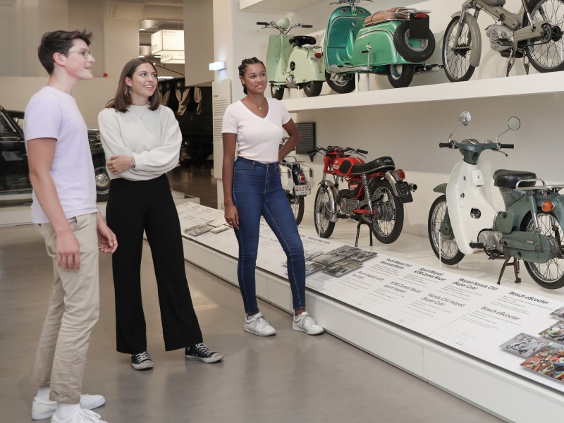 A small group of teenagers look at various mopeds and motorcycles in the exhibition "Mobility".: 