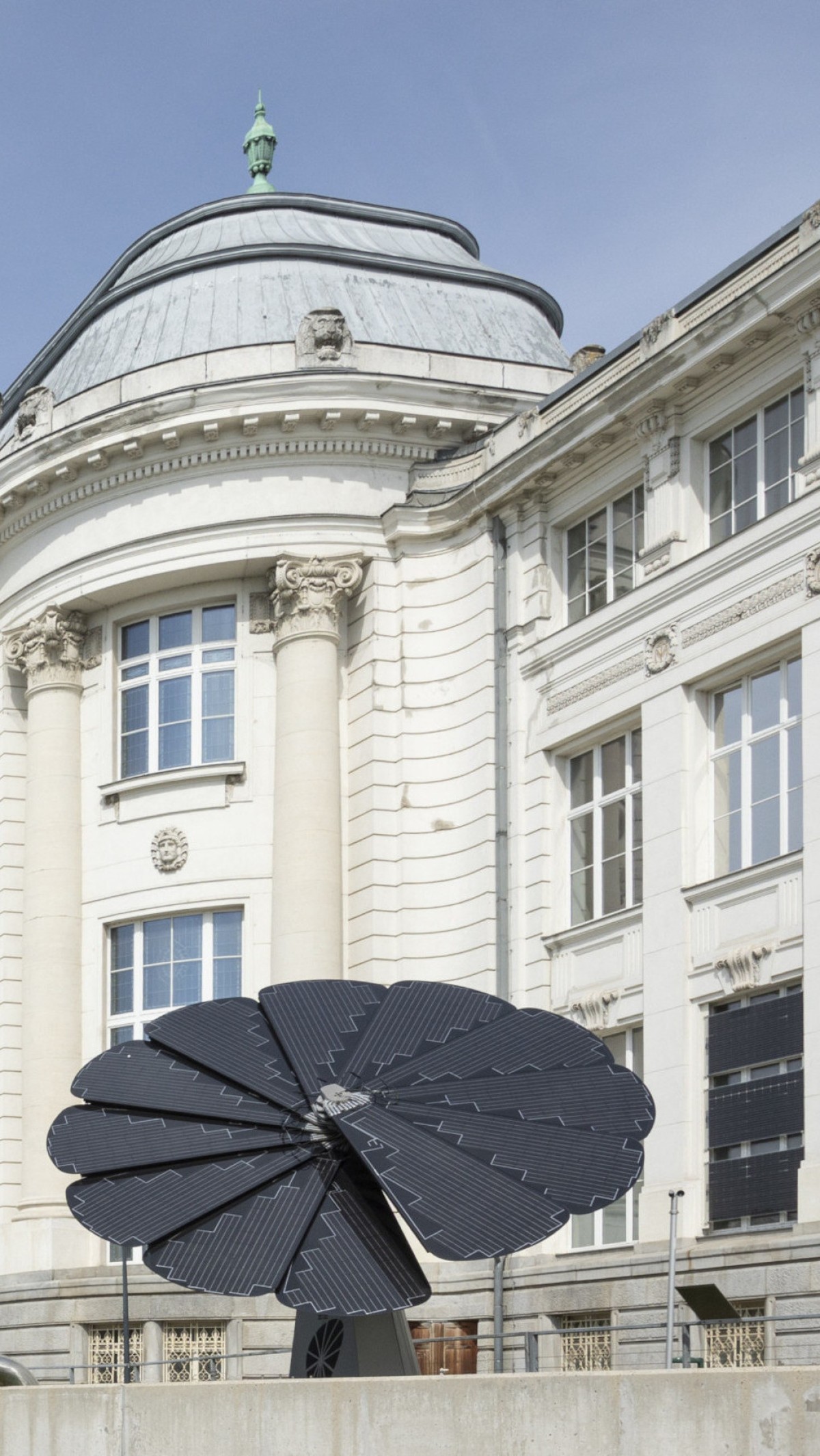 Eine fächerförmige Photovoltaik-Anlage in Form einer dunklen Blume (SmartFlower) steht auf dem Vorplatz des Technischen Museums Wien. Im Hintergrund ist die prachtvolle, weiße Architektur des Museums mit Säulen, Rundbogenfenstern und einer markanten Dachkuppel unter blauem Himmel zu sehen.: ... zentrales Anliegen des Museums