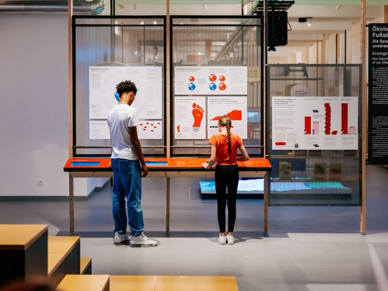 Zwei Personen, ein junger Mann und ein Mädchen, stehen vor einer interaktiven Ausstellung im Museum. Die Exponate hinter Glas und auf Tafeln zeigen Diagramme, Texte und Grafiken zum Thema ökologischer Fußabdruck und Umweltbewusstsein. Die Ausstellung scheint sich mit nachhaltigem Leben und Umweltschutz zu befassen. Der Raum ist modern gestaltet, mit hellen Wänden und Holzrahmenstrukturen.reen in einer Ausstellung mit informativen Tafeln und weiteren Exponaten im Hintergrund.