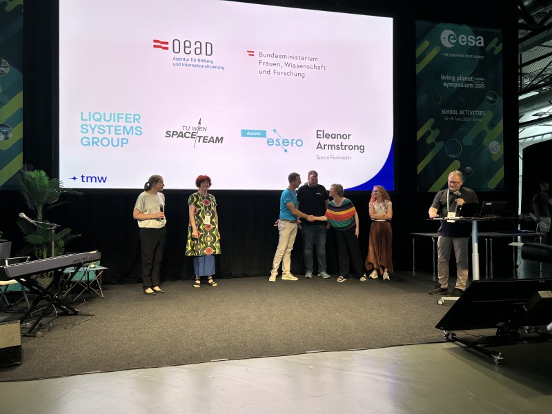 Personengruppe auf Bühne vor Leinwand mit Logos von Partnerorganisationen, eine Person spricht am Rednerpult.: Die Projektpartner:innen erzählen von der Wirkung, die die Teilnahme an „This is (not) rocket science!“ auf sie hat.