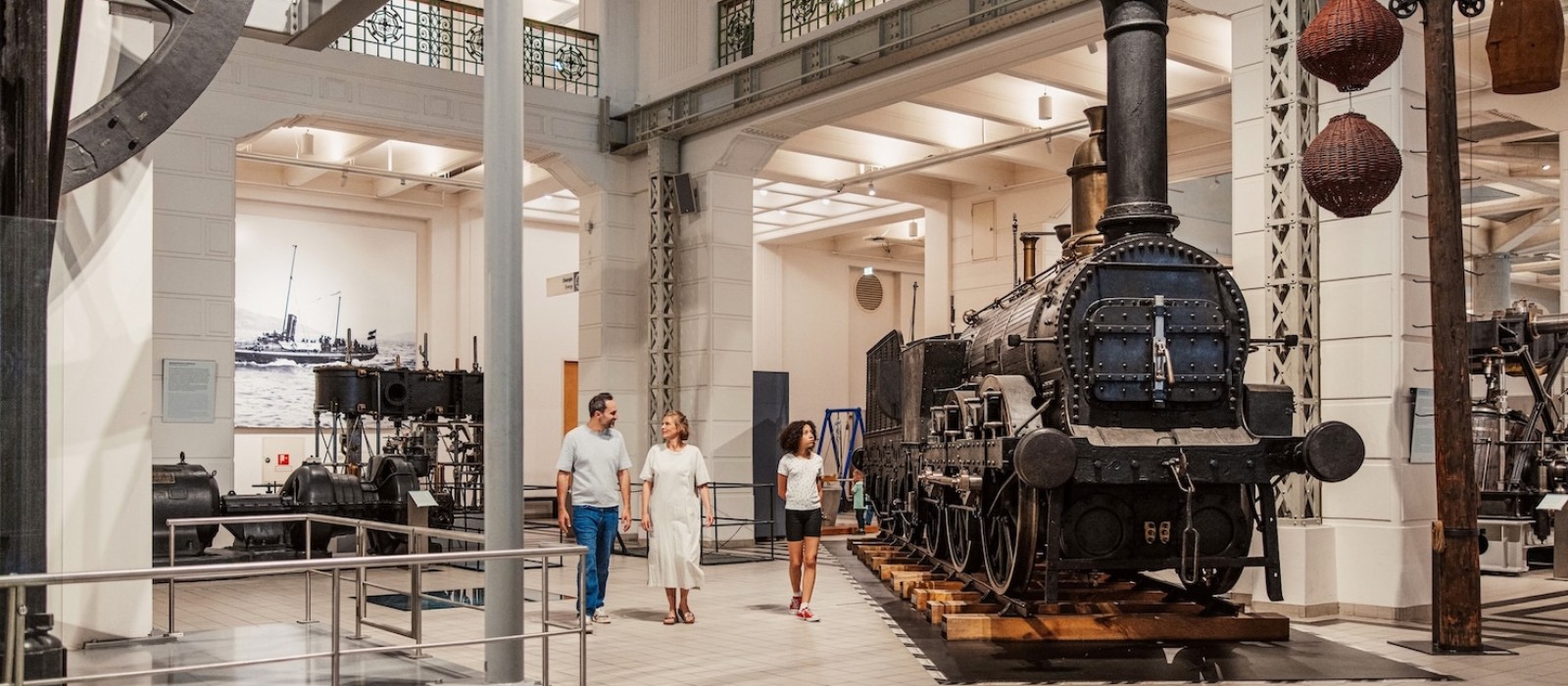 Drei Personen gehen durch eine helle Ausstellungshalle mit alten Maschinen und einer Dampflokomotive.: