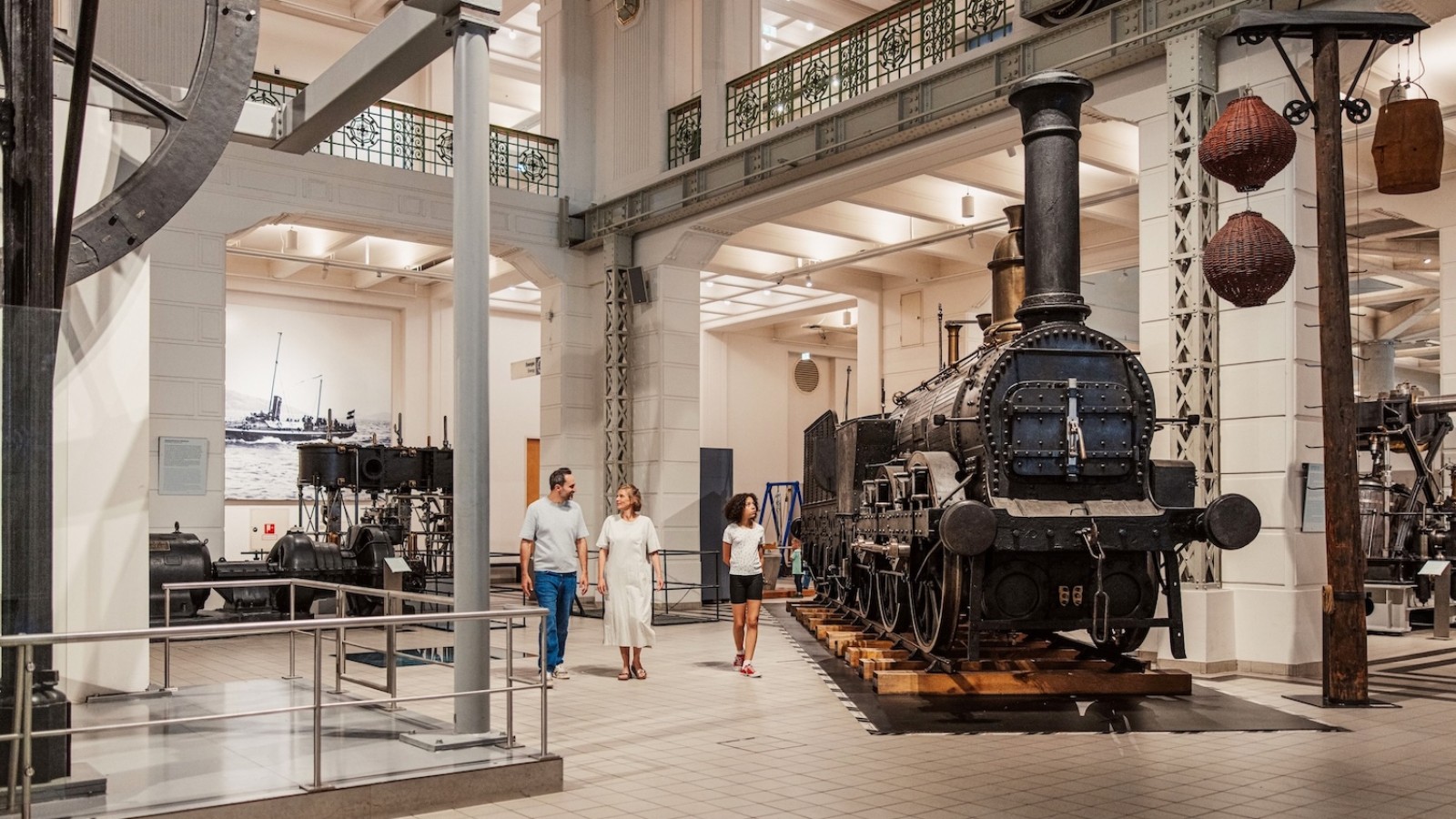Drei Personen gehen durch eine helle Ausstellungshalle mit alten Maschinen und einer Dampflokomotive.: