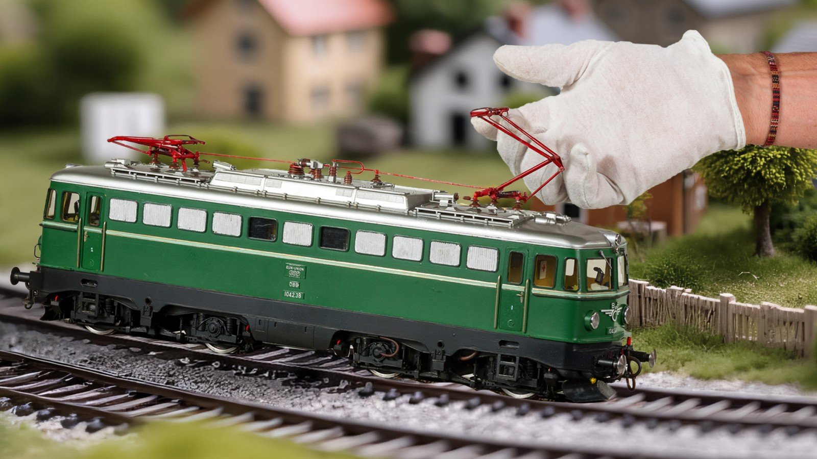 Eine Hand mit weißem Handschuh hebt den roten Stromabnehmer einer grünen Modell-Elektrolokomotive auf einem Gleis an; im Hintergrund stehen modellhafte Häuser und Bäume.: 