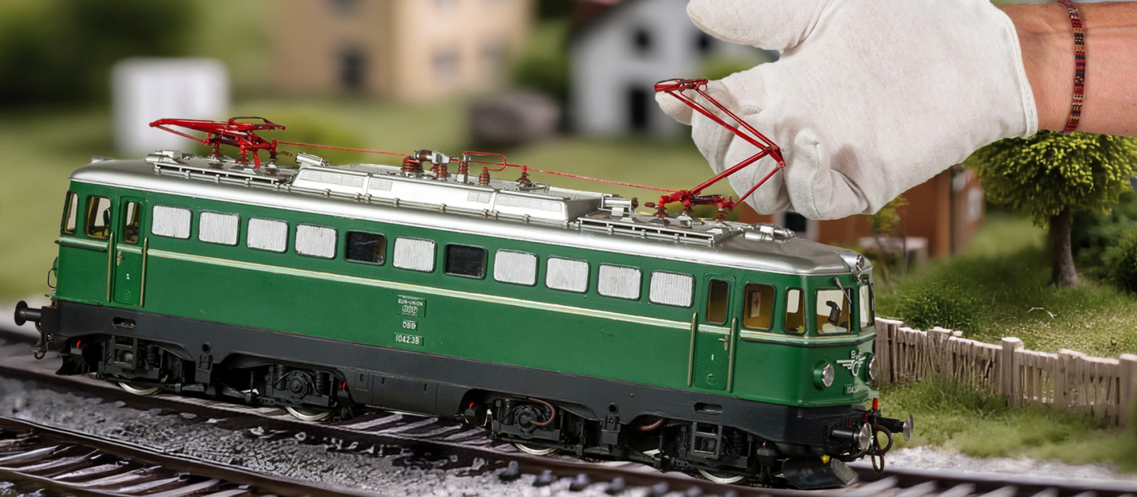 Eine Hand mit weißem Handschuh hebt den roten Stromabnehmer einer grünen Modell-Elektrolokomotive auf einem Gleis an; im Hintergrund stehen modellhafte Häuser und Bäume.: 