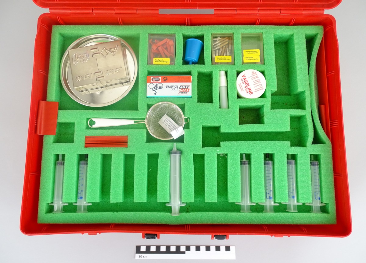 Geöffnetes rotes Werkzeugkoffer-Set mit grünem Schaumstoff-Einlage, das eine Metallplatte, Sieb, Spritzen, Kleinteile und eine Vaseline-Dose enthält.: CVK-Experimentierbox 31705 zum Thema „Luft“ (Inv.-Nr. 113.239)